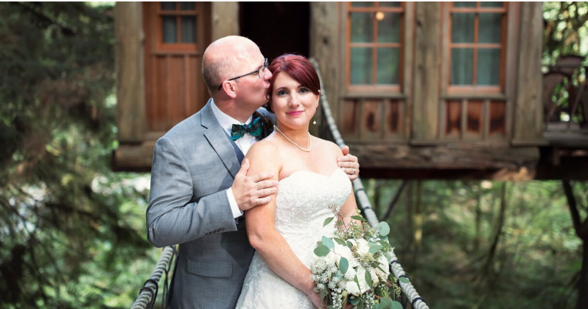 Couple in wedding attire standing in front of a treehouse.